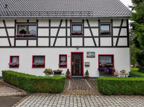 Ein hübsches Fachwerkhaus mit weißer Fassade und roten Fenstern. Vor dem Haus gibt es gepflegte Pflanzen und einen gepflasterten Weg.