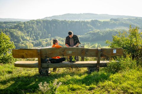 Zwei Personen sitzen auf einer Bank mit Blick auf eine grüne Landschaft. Die Umgebung ist sonnig und friedlich.