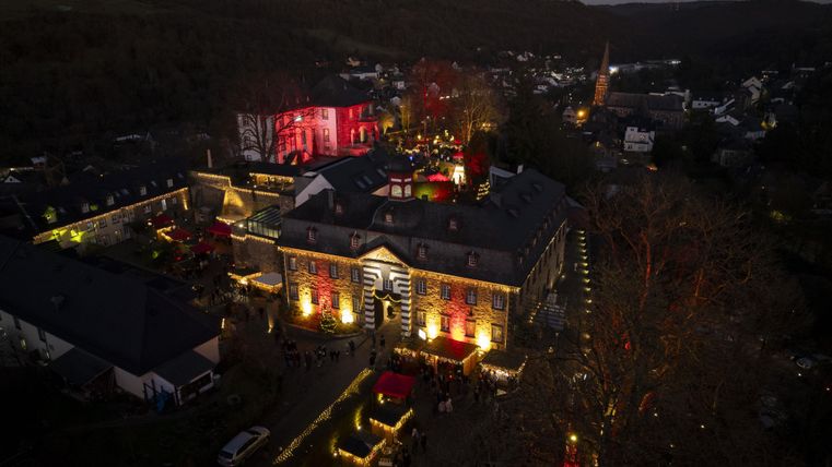 Eine festlich beleuchtete historische Gebäudeansicht bei Nacht. Rund um das Gebäude sind Menschen und bunte Lichter zu sehen.