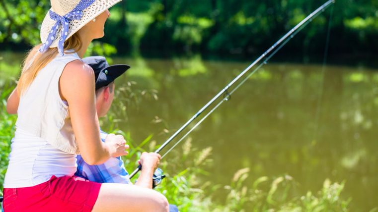 Two summery dressed people are sitting next to each other on the shore of a body of water, holding fishing lines in the water. Around them are meadows and forests.