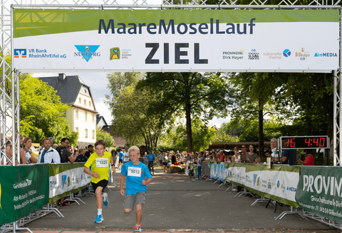 Deux garçons courent avec enthousiasme au-dessus de la ligne d'arrivée d'une course. En arrière-plan, des spectateurs et une bannière d'arrivée verte sont visibles.