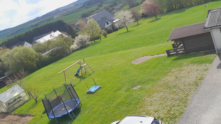 Een grote weide met een trampoline, een schommel en een kleine speeltuin. Op de achtergrond zijn bomen en enkele huizen te zien.