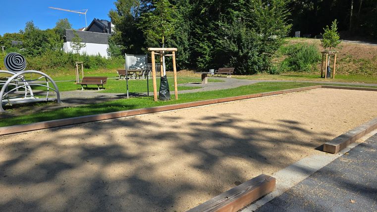 A child-friendly playground with a sand area and play equipment. In the background, green trees and seating are visible.