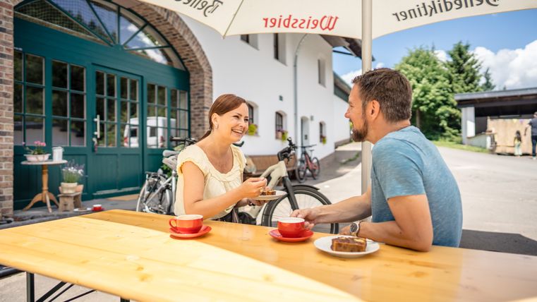 Un couple est assis à une table en bois à l'extérieur, il boit du café et mange du gâteau. En arrière-plan, on aperçoit des vélos et un bâtiment.