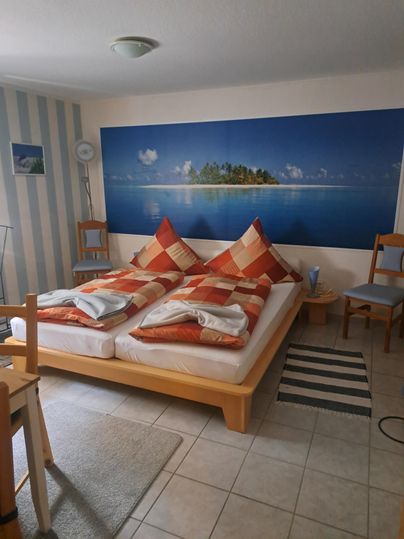 Een slaapkamer met een bed en kleurrijke kussens. Aan de muur hangt een schilderij van een tropisch eiland.