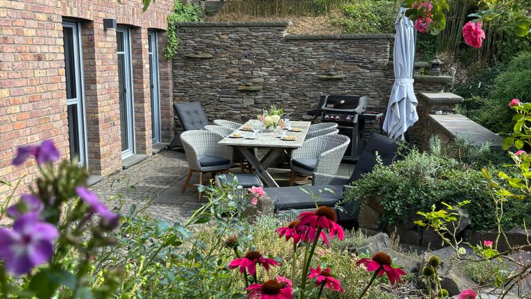 A beautiful garden with colorful flowers and a cozy seating area. In the background, there is a modern house with large windows.