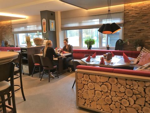 Een gezellige café-ruimte met rode banken en een houten decoratie. Twee vrouwen zitten aan de tafel en praten met elkaar.