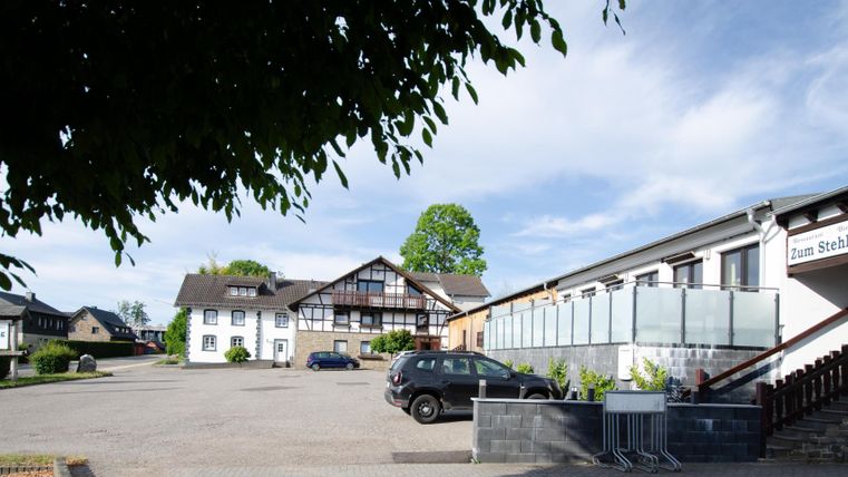 Een rustige parkeerplaats voor een hotel met meerdere gebouwen. De lucht is helder en zonnig, omringd door bomen.