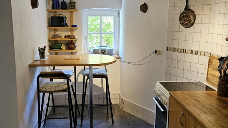 Eine gezellige keuken met een kleine tafel en hoge stoelen. Aan de muur hangen open planken met servies en keukenspullen.
