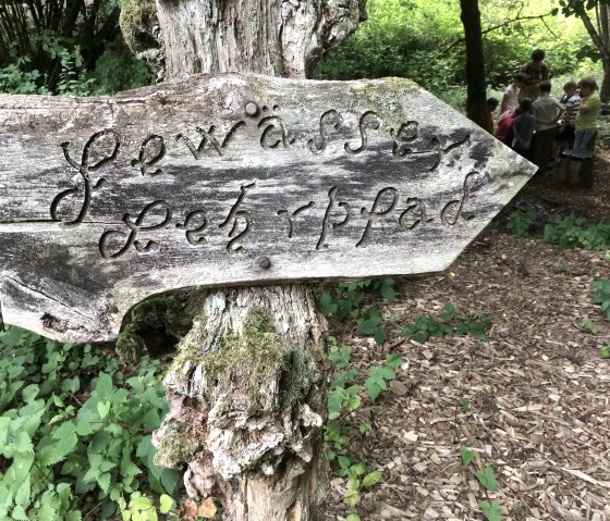 Ein altes Holzschild mit der Aufschrift 'Gewässer Lehrpfad' zeigt den Weg in einem bewaldeten Gebiet. Im Hintergrund sind Menschen zu sehen., © TI Bitburger Land