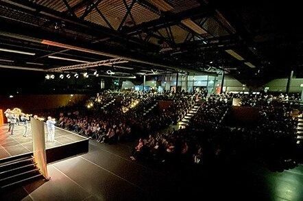 Een groot publiek zit in een moderne locatie. Op het podium staan meerdere mensen die een presentatie geven.