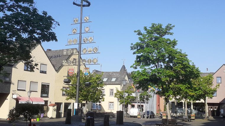 La place Graf-Mirbach à Hillesheim avec quelques magasins et des maisons résidentielles tout autour et un arbre des corporations au milieu.