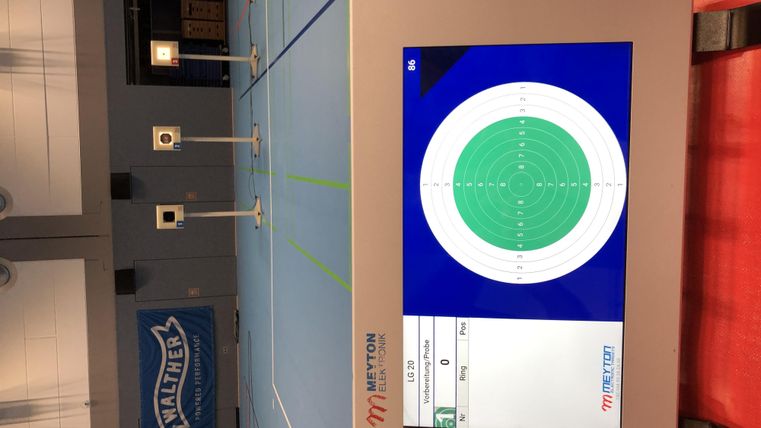 Ein digitaler Bildschirm zeigt ein Ziel mit einem grünen Bereich in einer Schießanlage. Im Hintergrund sind mehrere Zielscheiben zu sehen.