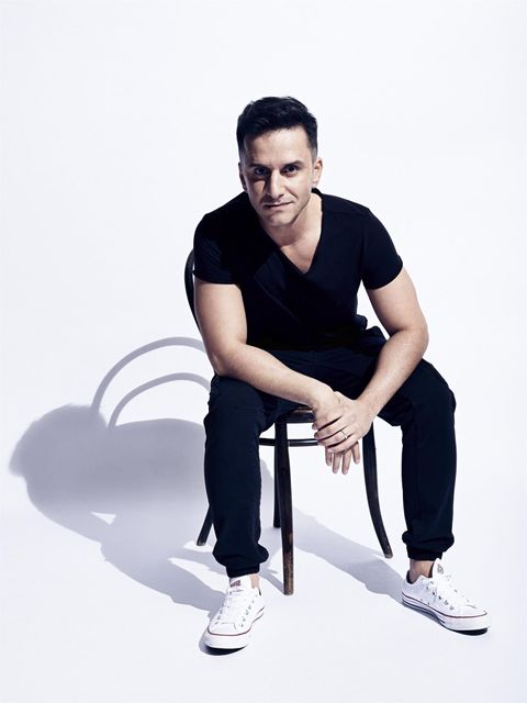 Un homme est assis sur une chaise et porte un t-shirt noir ainsi que des pantalons noirs. L'arrière-plan est clair et neutre, ce qui attire l'attention sur la personne.