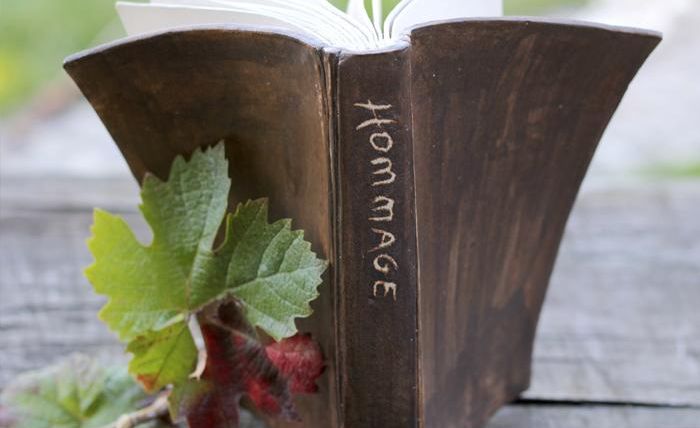 A stylish book with the engraving "HOMMAGE" on the cover stands on a wooden base. Surrounding it are vine leaves and grapes.