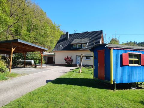 Ein schönes Haus mit einem Carport und einem bunten Bauwagen im Garten. Umgeben von grünen Bäumen und klarem Himmel.