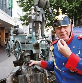A man in a historical uniform stands next to a fountain figure. In the background, additional figures and a cart can be seen.