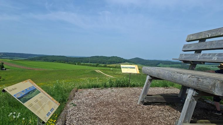 Un banc en bois se trouve sur un point de vue avec un champ vert et un ciel bleu en arrière-plan. À côté du banc, des panneaux d'information sont installés.