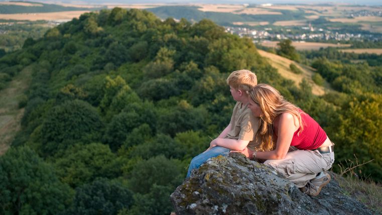 Zwei Personen sitzen auf einem Felsen mit Blick auf eine bewaldete Landschaft.