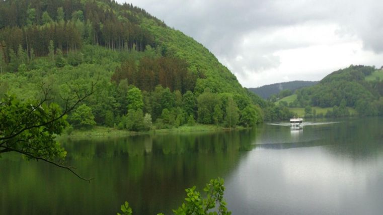 Un lac paisible entouré de collines vertes et d'arbres. En arrière-plan, un petit bateau est visible sur l'eau.