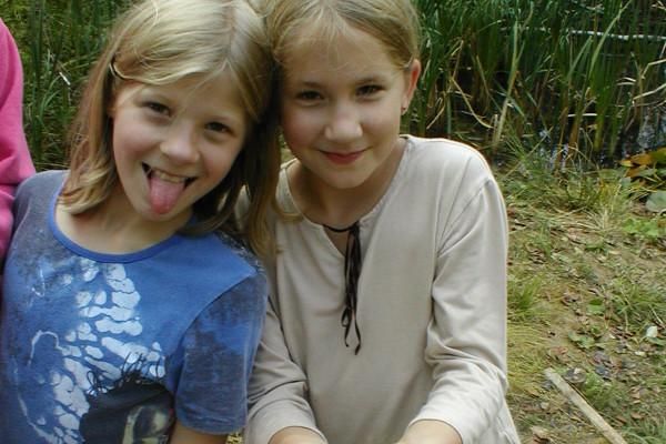 Zwei lächelnde Mädchen stehen im Freien. Sie posieren fröhlich und halten etwas in den Händen.