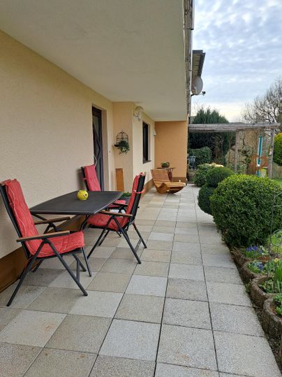 Een gezellige buitenruimte met een tafel en stoelen op een stenen terras. Groene struiken en planten geven de omgeving een uitnodigende sfeer.