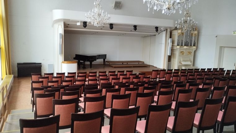 Ein großer Saal mit vielen roten Stühlen und einer Bühne. Auf der Bühne steht ein Klavier und im Hintergrund sind kunstvolle Elemente zu sehen.