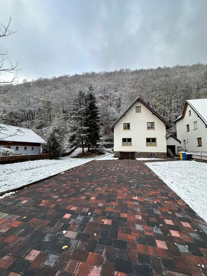 A snowy path leads to a white house. In the background, snow-covered trees can be seen.