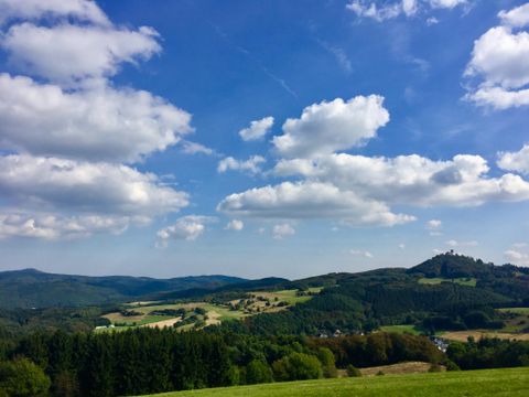 Landschaft mit Burgruine Nürburg auf einem Hügel, umgeben von Wäldern und Feldern unter blauem Himmel mit Wolken.