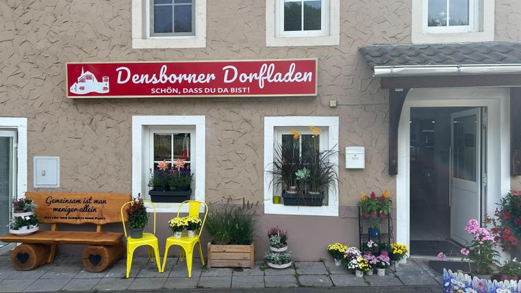 A small village shop named "Densborner Dorfladen" with a bright facade. Colorful flowers and a wooden bench for sitting are in front of the shop.