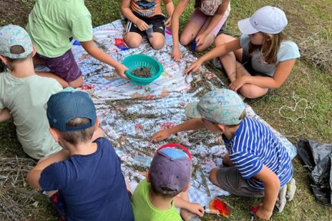 A group of children is sitting on a cloth and playing with various materials. They are using a bowl and appear focused and happy.