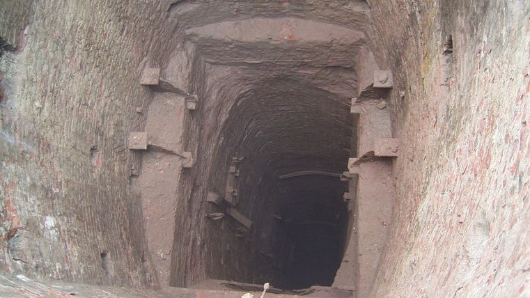 A deep shaft with steep walls and a barrel-shaped background. The floor is covered with dust and small plants.