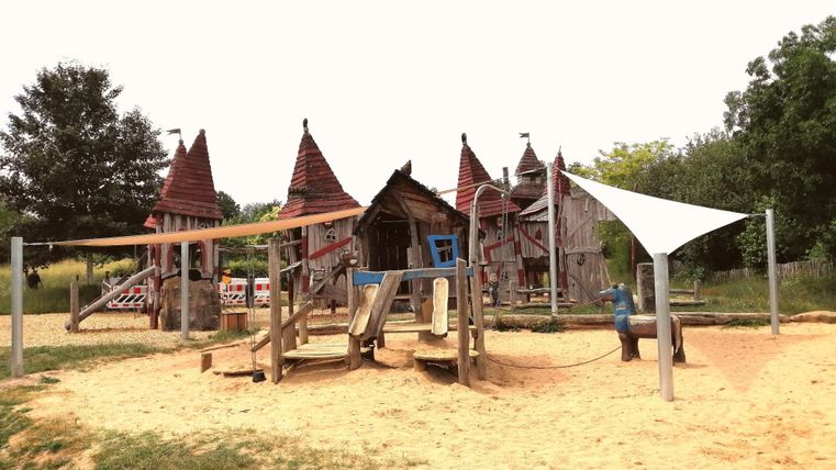 Ein Spielplatz mit einer Holzkonstruktion im Hintergrund und mehreren Spielgeräten. Sandboden und einige Bäume umgeben die Fläche.