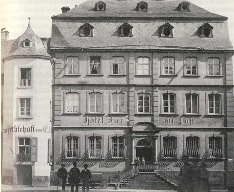 Een historisch gebouw met meerdere ramen en een ingang is te zien. Voor het hotel staan enkele personen, en de tekst "Hotel Fietz zur Post" is duidelijk te herkennen.