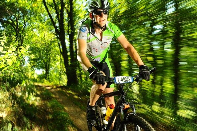 Ein Mountainbiker fährt schnell durch einen Waldweg, umgeben von grünen Bäumen. Er trägt einen Helm und eine grün-weiße Sportkleidung.