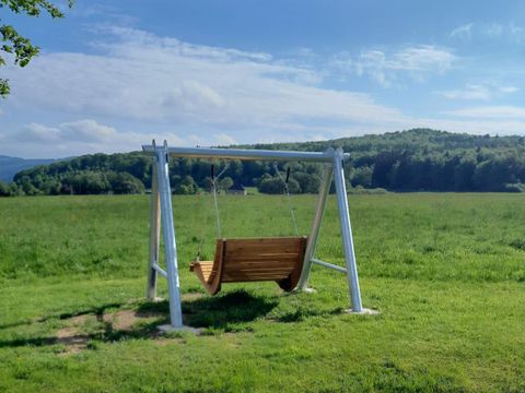 A swing stands on a meadow with a view of gentle hills. The sky is clear and blue, perfect for a relaxing day outdoors.