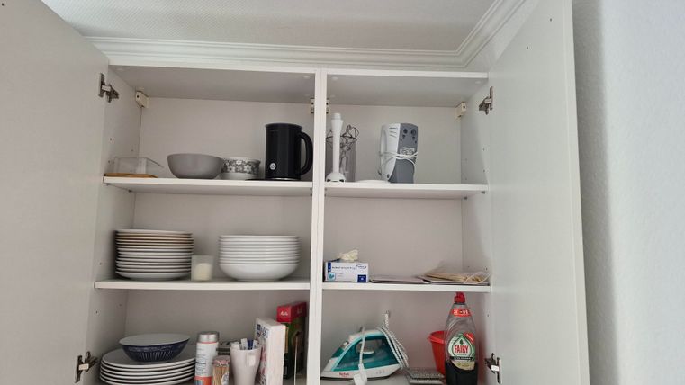 Een keukenkast met meerdere planken die servies, kopjes en keukenapparaten bevatten. Enkele voorwerpen zoals een koffiezetapparaat en een strijkijzer zijn ook zichtbaar.