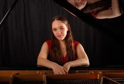 Ein junges Mädchen sitzt an einem Klavier und lächelt in die Kamera. Sie trägt ein rotes Oberteil und hat lange, braune Haare.