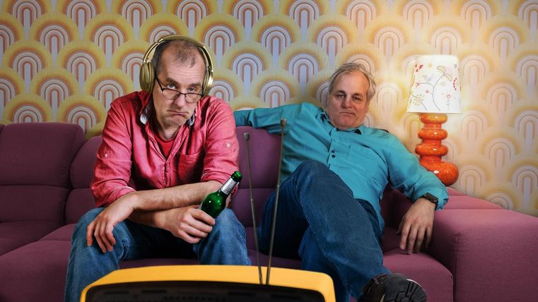 Zwei Männer sitzen auf einer lila Couch und schauen fern. Einer trägt Kopfhörer und hält eine Flasche Bier, während der andere entspannt aussieht.