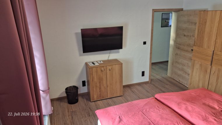 A modern bedroom with a double bed and a television on the wall. Next to the bed is a wooden piece of furniture and a trash can is visible.