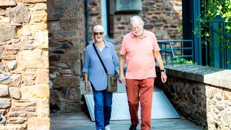 Ein älteres Paar geht nebeneinander auf einem Weg aus Stein. Die beiden sind auf der Burg Hengebach. Um sie herum sind hohe Mauern aus braunem Stein. Es ist ein sonniger Tag.