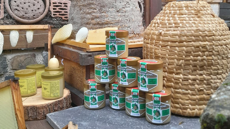Op de voorgrond staan 8 glazen honing op elkaar gestapeld, naast hen staat een andere soort honing, op de achtergrond staat een bijenkorf van stro en verdere decoratieartikelen.