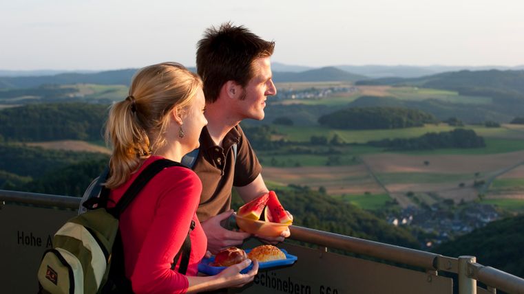 A couple are standing on a lookout tower and gazing into the distance. They are holding snacks in their hands. A hilly landscape can be seen in the background.