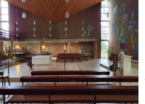 Eine moderne Kirche mit einem hellen Innenraum und einer auffälligen Holzdecke. Im Vordergrund stehen Bänke, und im Hintergrund ist ein Altar sowie bunte Wandmalereien zu sehen.