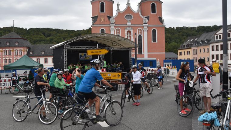 A group of cyclists gathers in front of a historic church. In the background, buildings and a stage can be seen.