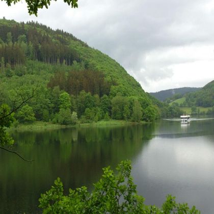 Een rustige meer omringd door groene heuvels en bomen. Op de achtergrond is een klein bootje te zien.