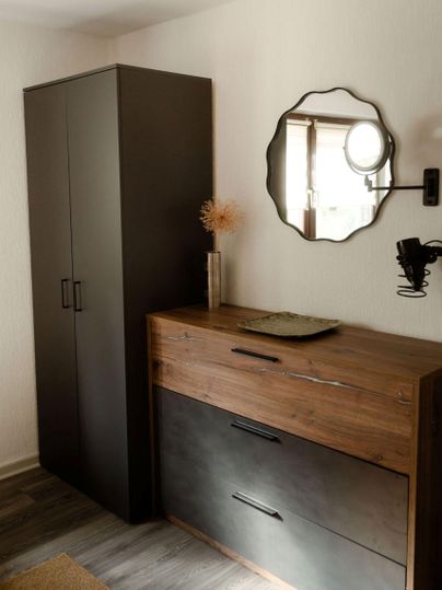 A modern bedroom with a black wardrobe and a wooden dresser. A decorative mirror hangs on the wall.