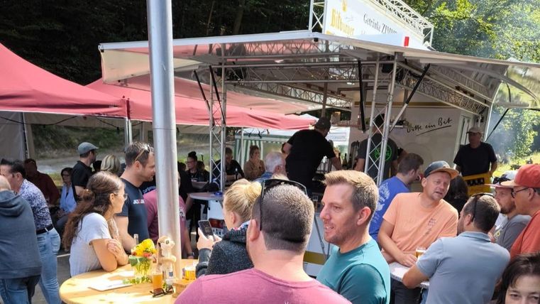 A social event with many people standing at a food stall. There are chairs, tables, and sun umbrellas outside.