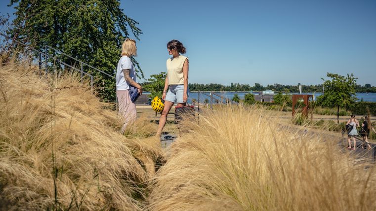 Two people with helmets in their hands are standing on a staircase in the foreground. On the right of the picture, a path leads through the Seepark Zülpich, on which two other people are walking. A lake surrounded by trees can be seen in the background.