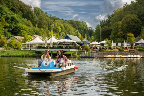 Deux femmes naviguent sur un pédalo sur un lac calme. En arrière-plan, on peut voir des arbres et un terrain de loisirs confortable.